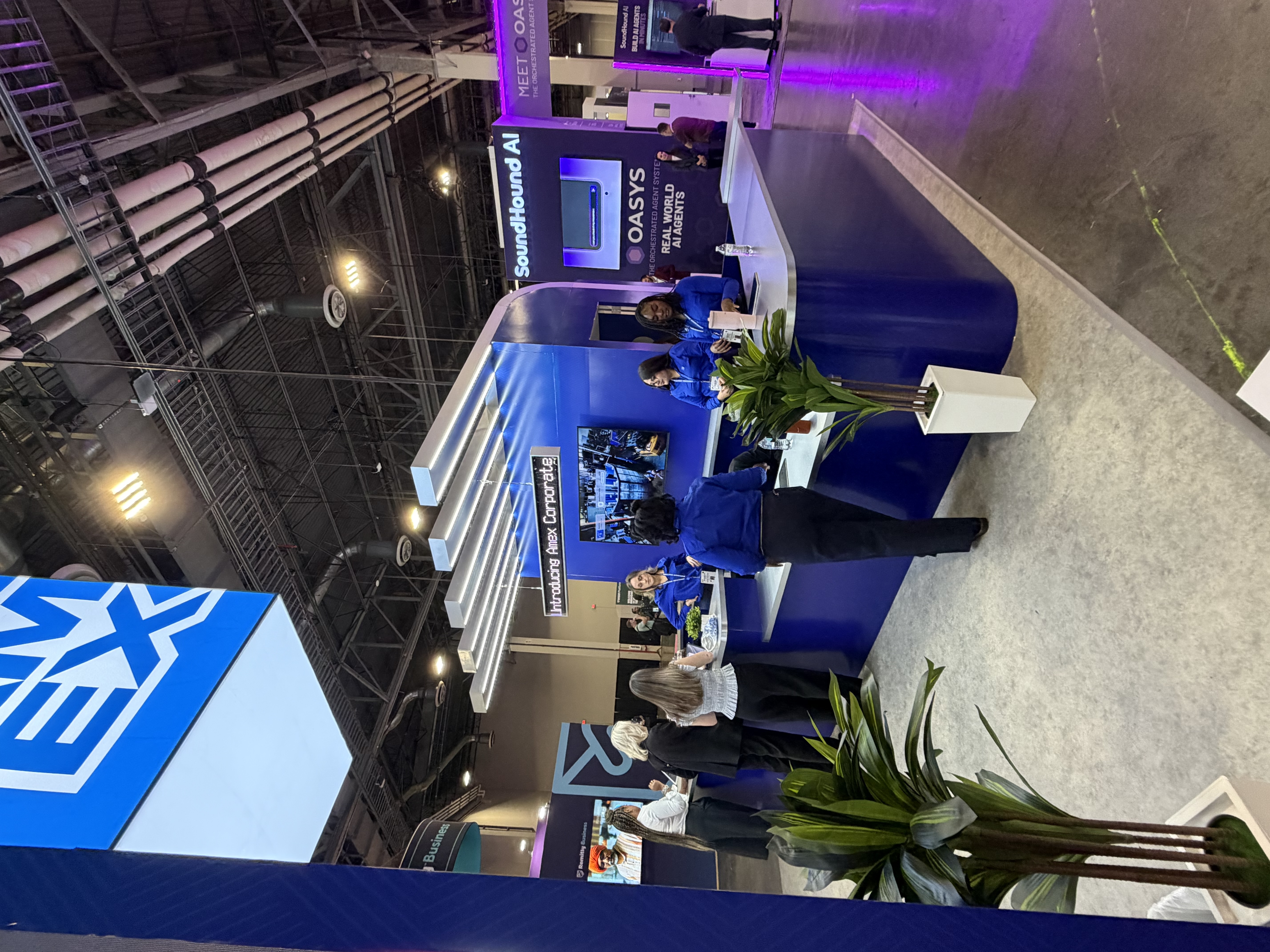 American Express exhibit viewed from the opposite aisle, with curved blue counters, illuminated overhead beams and staff at Money20/20 2026